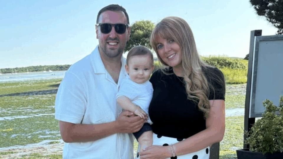 Leah and George standing outdoors, holding baby George and smiling at the camera