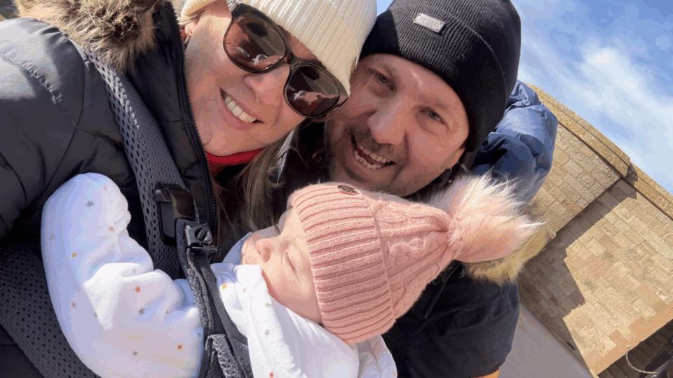 Helen wearing a hat and sunglasses, standing outdoors next to Stephen in a black hat, holding Faith in sling.