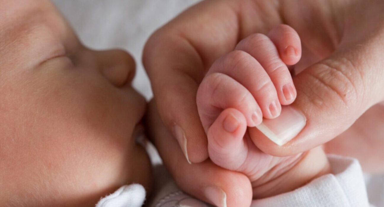 A baby is sleeping and their parent is holding their hand. The baby is gripping their parent's thumb,