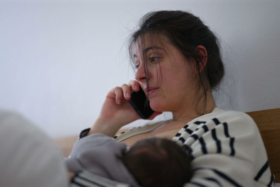 Mother multitasking while her baby sleeps peacefully in her arms, speaking on the phone with a thoughtful expression. the quiet balance between caring for a sleeping newborn and handling daily responsibilities