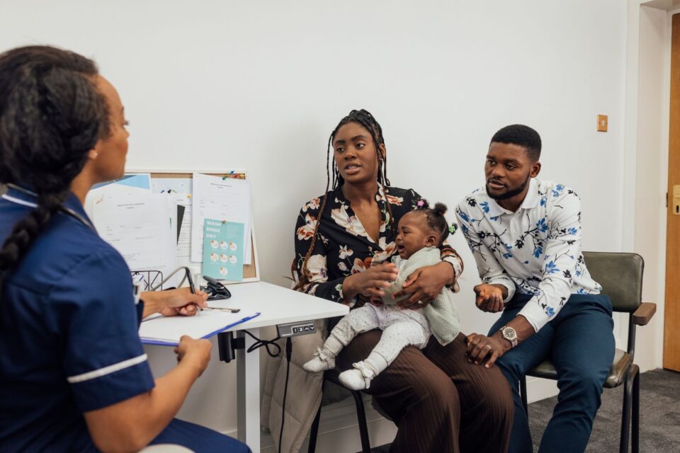 A young couple visiting a medical clinic in Newcastle upon Tyne, England with their baby daughter for a check up. They are sitting in a doctor's office while the nurse listens to their concerns about their baby, who is crying.