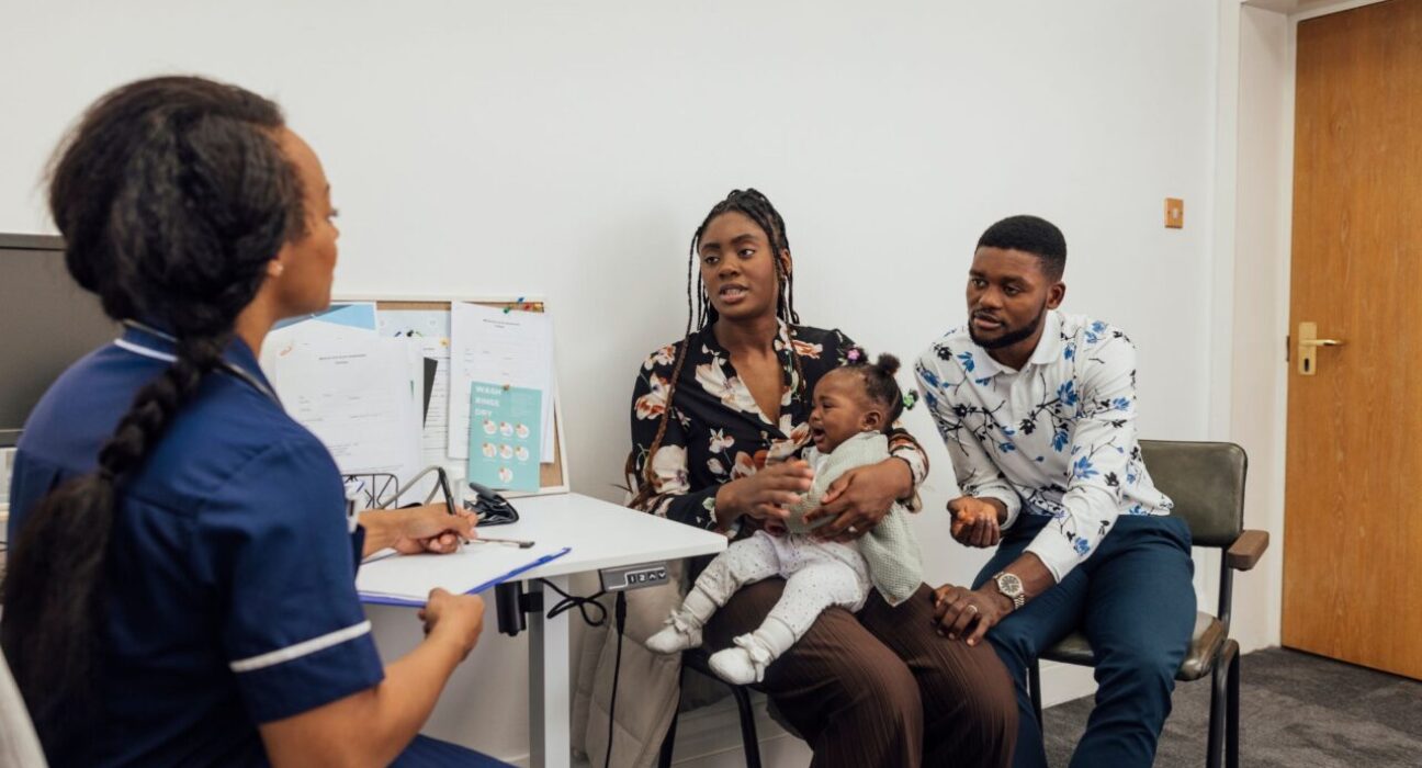 A young couple visiting a medical clinic in Newcastle upon Tyne, England with their baby daughter for a check up. They are sitting in a doctor's office while the nurse listens to their concerns about their baby, who is crying.