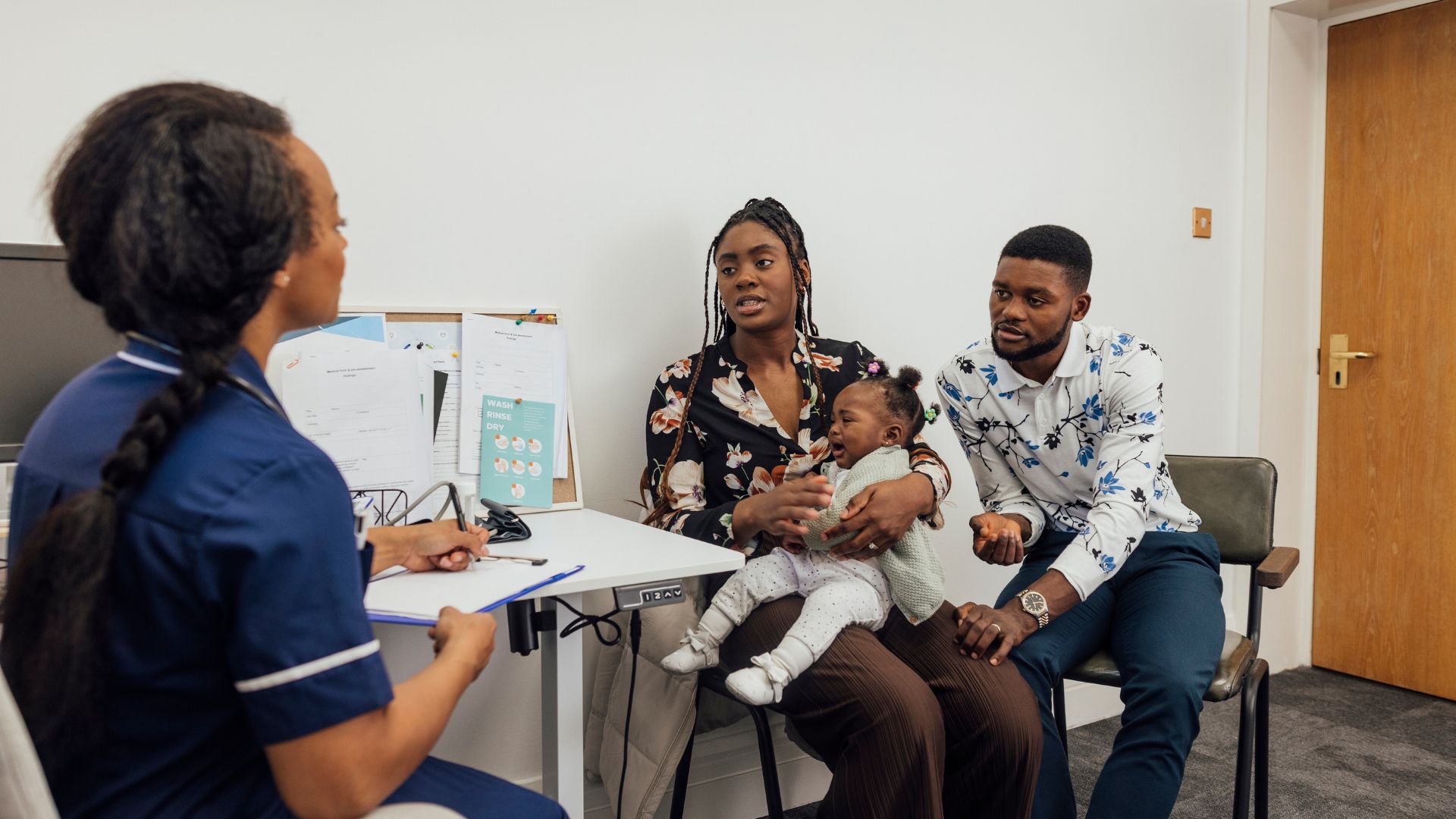 A young couple visiting a medical clinic in Newcastle upon Tyne, England with their baby daughter for a check up. They are sitting in a doctor's office while the nurse listens to their concerns about their baby, who is crying.