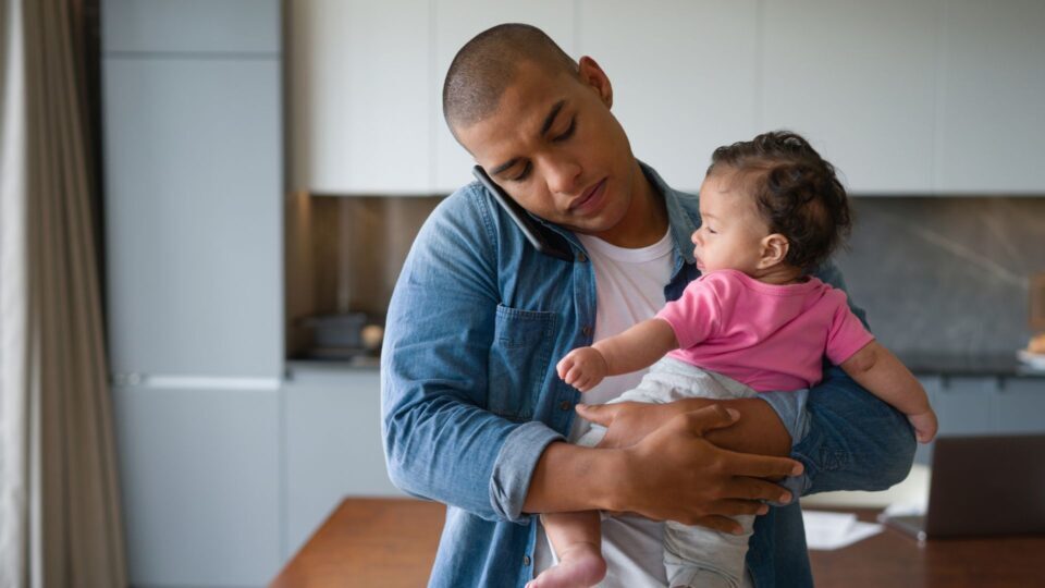 Busy Latin American father multi-tasking at home carrying the baby while talking on the phone