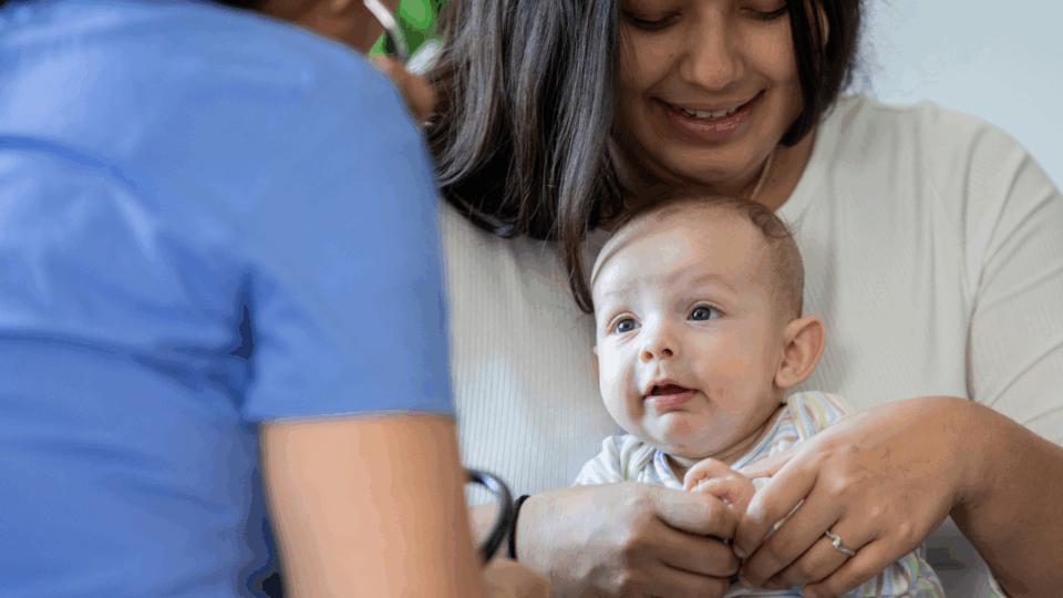 A baby is sat on her mum's knee, facing a health professional. The baby is smiling.