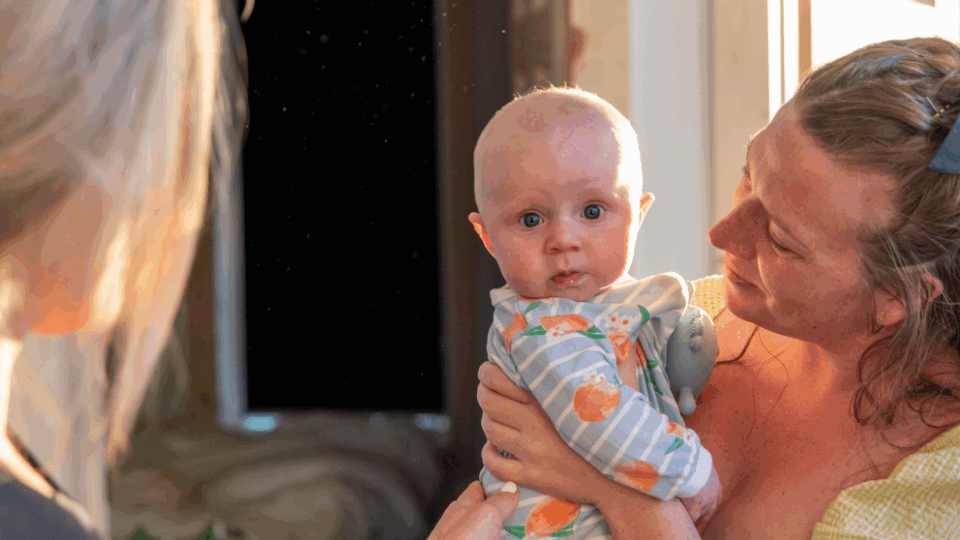 Lady holding a baby, and another lady standing in front of her with her hand on the baby's back.