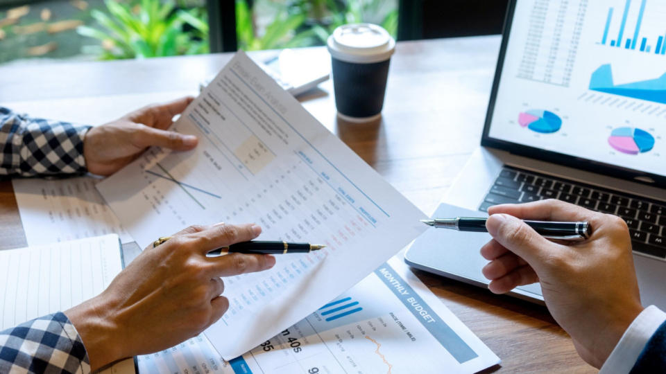 People's hands holding pens, inspecting graphs and data on sheets of paper.