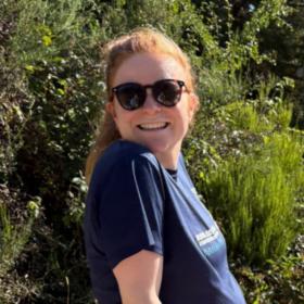Chloe outdoors wearing sunglasses on a sunny day, smiling at the camera.