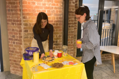 Two woman stand by a table which has a yellow table cloth on, refreshements and leaflets.