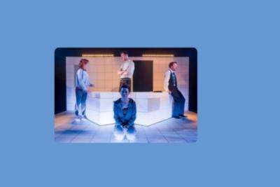 A photo of a theatre performance. There are four people stood on stage, all facing in different directions.