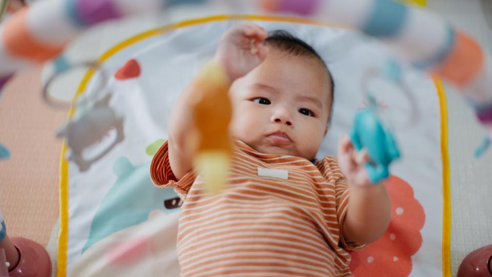 A baby is laying on their back playing with toys dangling above. They are wearing an orange and white stripey baby grow.
