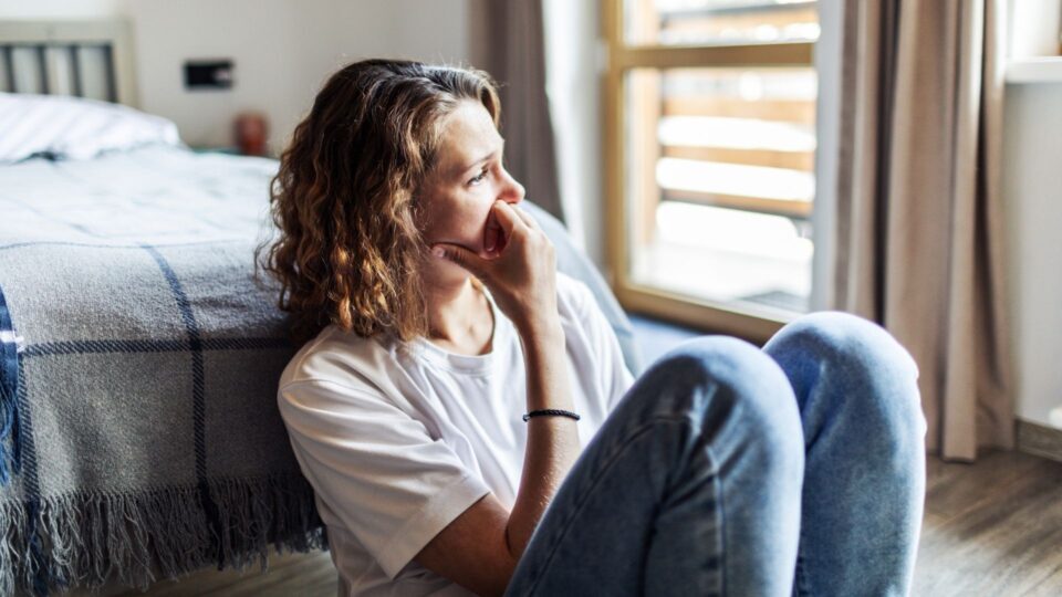A woman is sat against the foot of the bed, she looks worried and is looking out of the window. Her hand is rested against her face.
