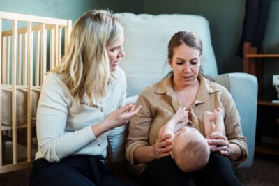 A mum is looking down at her baby in her knee. A health visitor is next to them, talking to the mother. A cot is in the background.