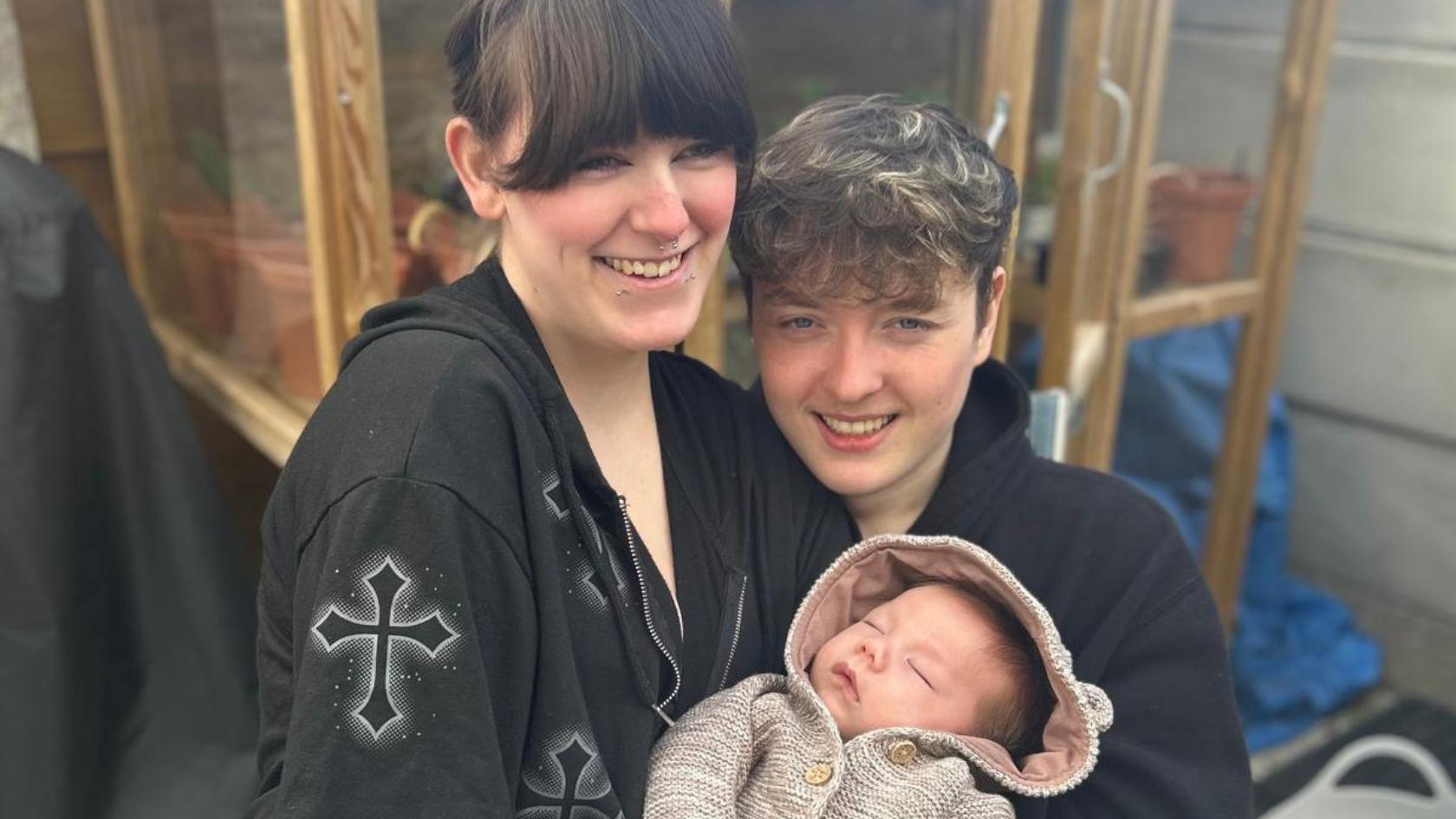 Lilly and James smiling at the camera, holding Theo.