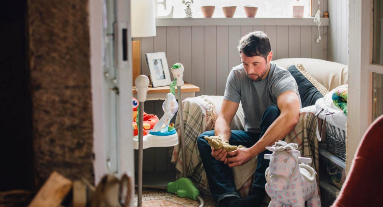 Man sitting on an armchair in a child's bedroom amongst toys, looking solemnly at a teddy bear in his hands.
