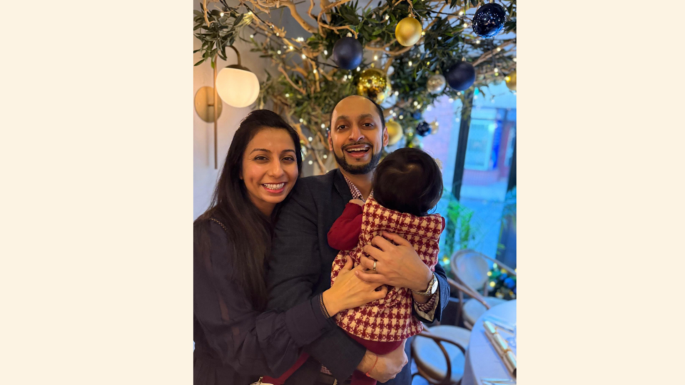 Harpal, Vishal and Lara are stood together. Vishal is holding Lara, with lara facing his chest and smiling widely and openly and Harpreet smiles beside him. They are stood infront of a bauble and tree display.