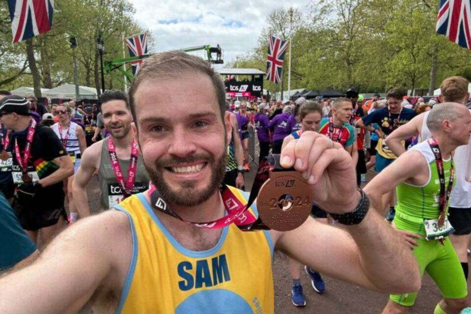 Man standing in crowd of runners smiles to camera and holds up a London marathon medal