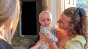A mum with red, curly hair and fair skin is holding her baby who is looking directly at us. The baby is also fair skinned and has a stripy baby grow on.
