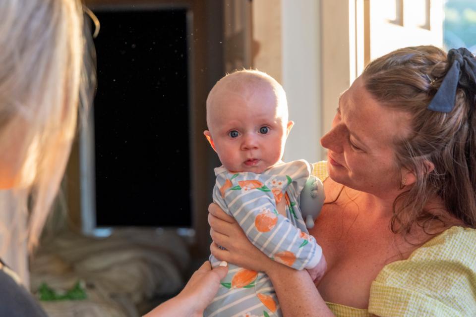 A mum with red, curly hair and fair skin is holding her baby who is looking directly at us. The baby is also fair skinned and has a stripy baby grow on.