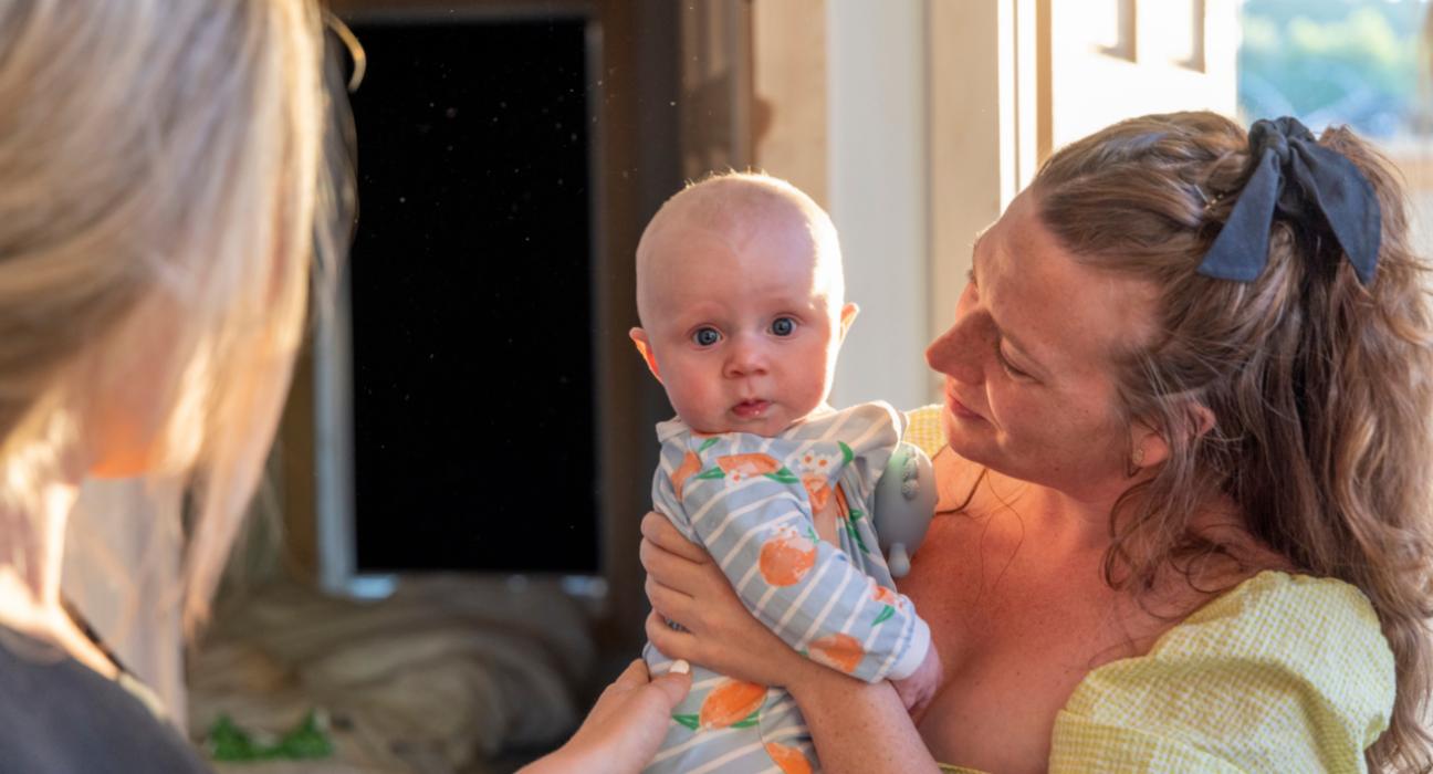 A mum with red, curly hair and fair skin is holding her baby who is looking directly at us. The baby is also fair skinned and has a stripy baby grow on.