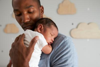 A Black dad is stood holding his baby on his chest, the baby is sleeping. The dad is looking down at the baby and appears to be settling them.