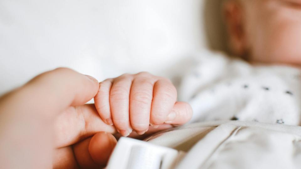 A fair baby's hand wraps around their parent's index finger, they are on white bedding
