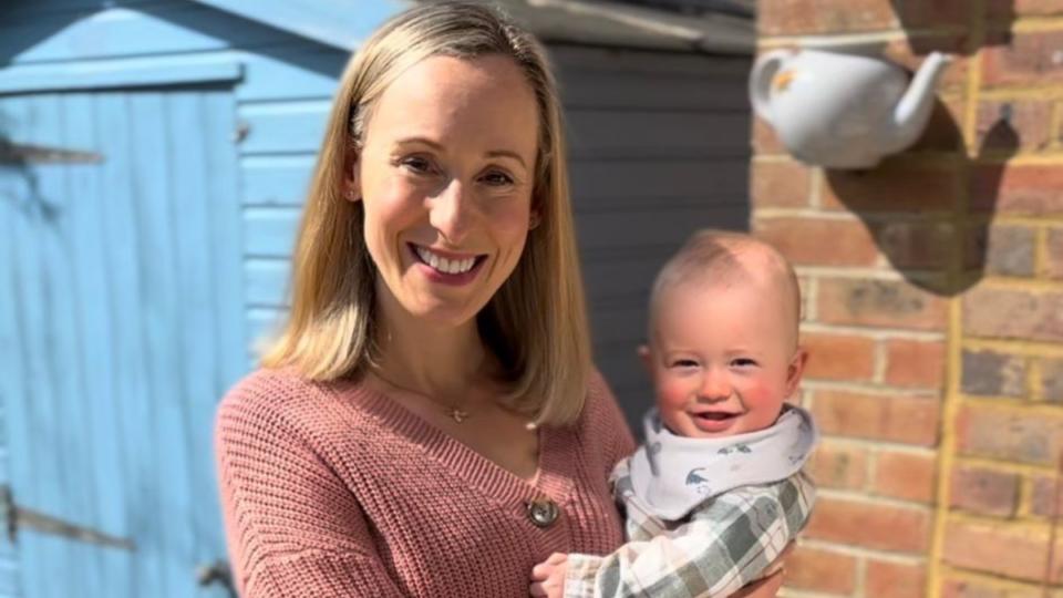 Laura standing outside holding her baby, smiling.