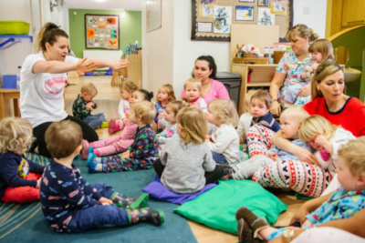 Children wearing pyjamas sit on the carpet in their nursery, watching their teacher