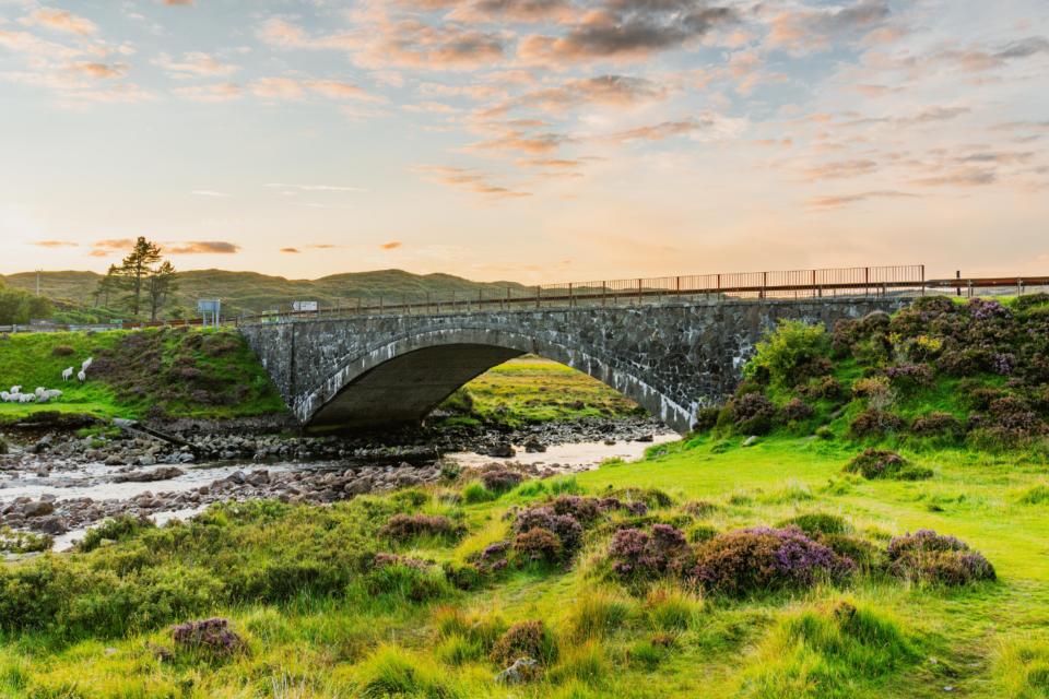 A shot of Scottish landscape. A stone bridge over a stream is surrounded by rolling, vivid green hills and moorlands.