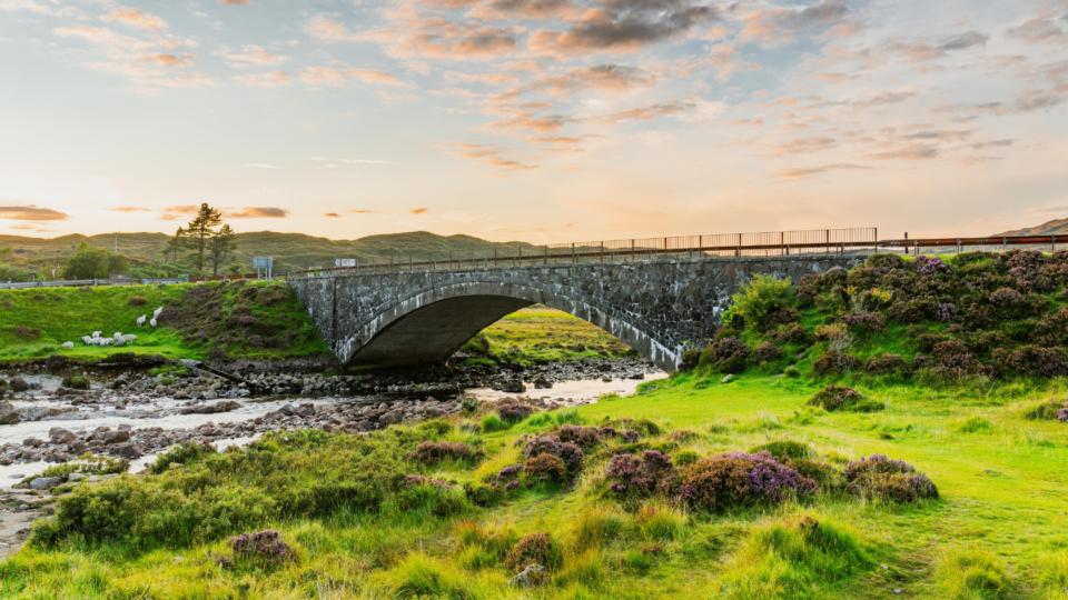 A shot of Scottish landscape. A stone bridge over a stream is surrounded by rolling, vivid green hills and moorlands.