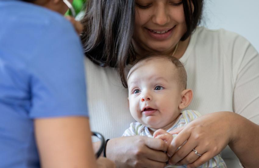 A baby is sat on her mum's knee, facing a health professional. The baby is smiling.