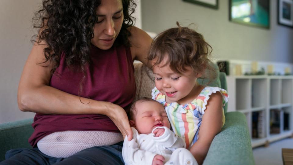 A woman sits on the sofa at home and helps her cute toddler daughter hold her baby brother for the first time. The big sister appears excited and proud.