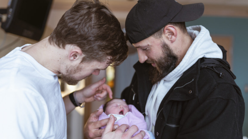 Two men holding a baby wrapped in a pink blanket.