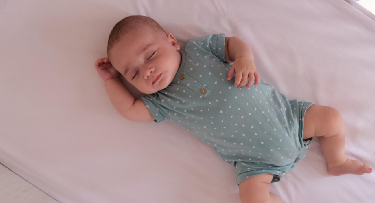 A baby sleeping safely on their back in an empty cot. They have one arm by their head, and another on their stomach. They are wearing a blue, polka-dot baby-grow.