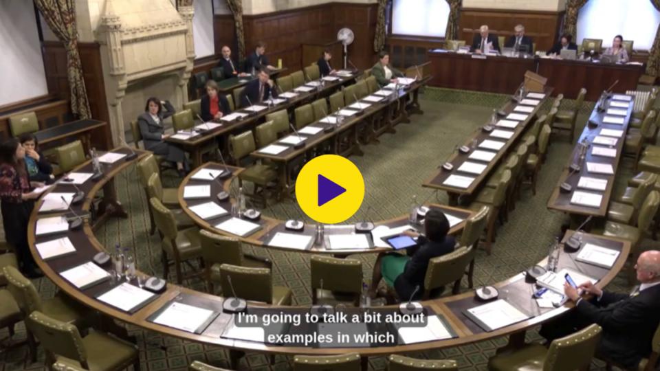 Screengrab of a video clip set at Westminster. There are long, curved tables and a handful of people sat at the table. There is a play icon in the middle.