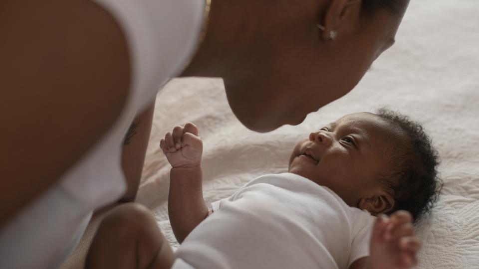 An infant African boy lays on a bed looking up at his mother. The young mother is leaning in close to his face as she looks down at him tenderly. They are both dressed casually.