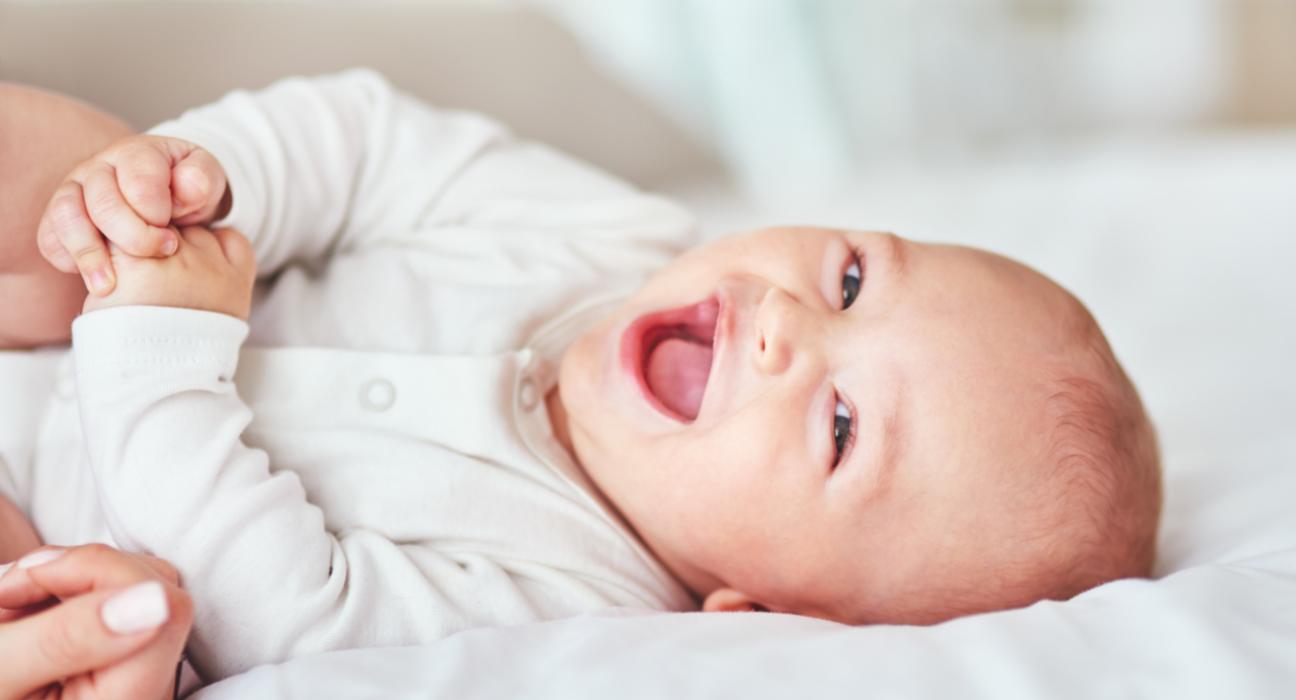 Baby is laying down smiling and giggling, grasping their hands together. They have light skin and wear a white babygrow.