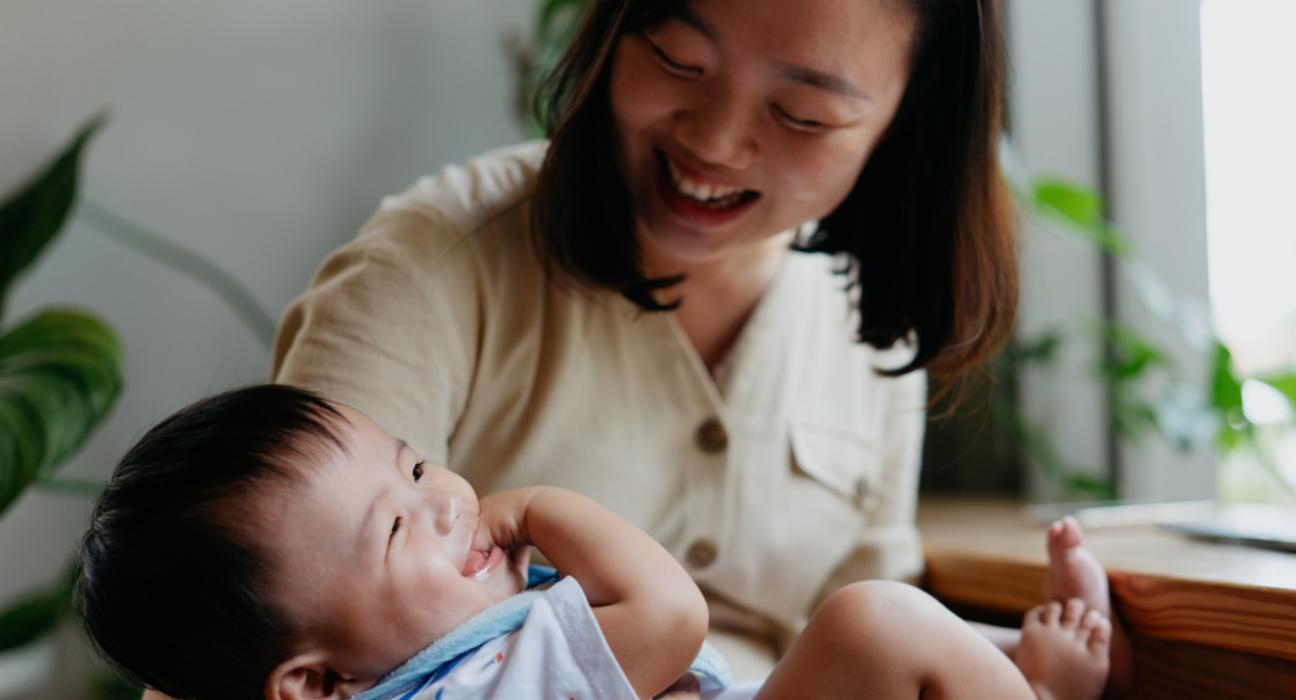 A mum is holding her baby in her arms. The baby is laughing and giggling with their hand in their mouth, and the mum is looking down at the baby, laughing too,