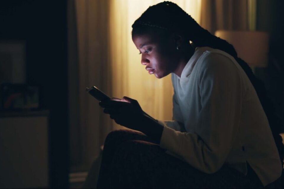 A black woman is sitting in a dark room, with the light from her phone shining on her face. She is crouched at the end of her bed. She looks sad, as though she's been crying.