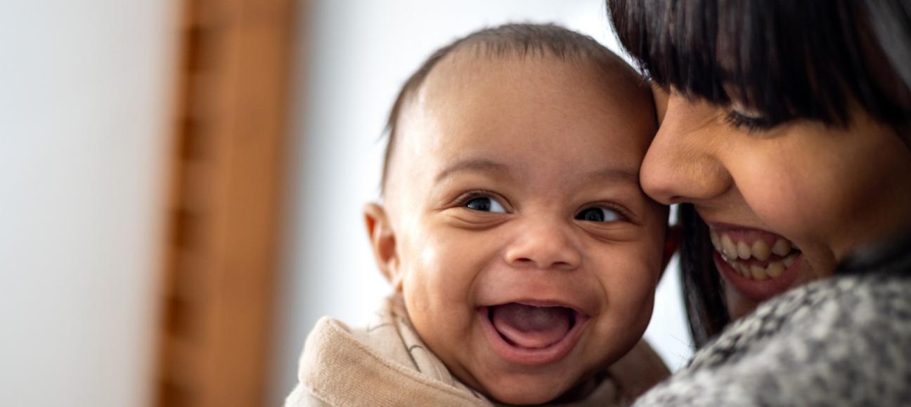 A mum is holding her baby close to her and laughing, and the baby is also laughing, looking to the right of the camera.