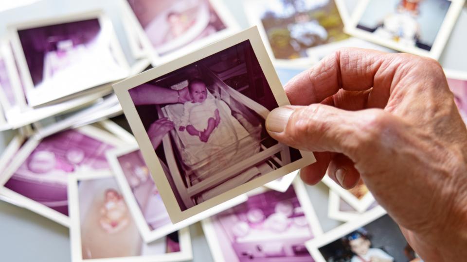 An older hand holding a photo of a baby. The photo is polaroid, looks dated and is of a baby in hospital after birth. There are blurred photos scattered in the background.