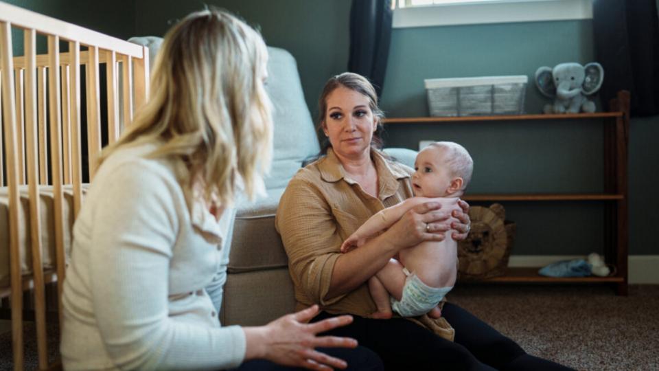 Parent holding her baby who is wearing a nappy, sitting on the floor talking to another lady, who is leaning on the cot.