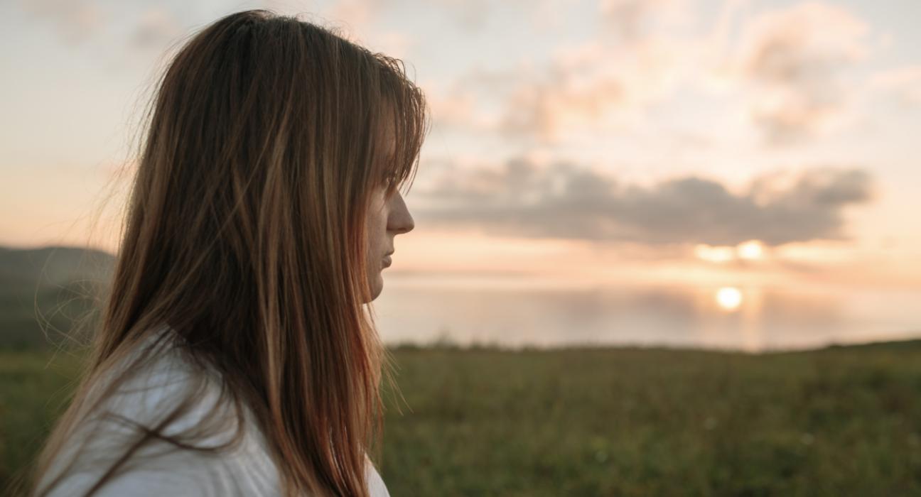 A woman is turned so you can see the side of her face. She is looking out in the distance, and seems empty.