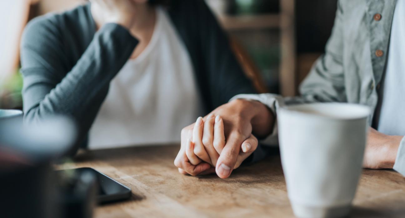 Two people are holding hands supportively. There is a phone and mug on the table.