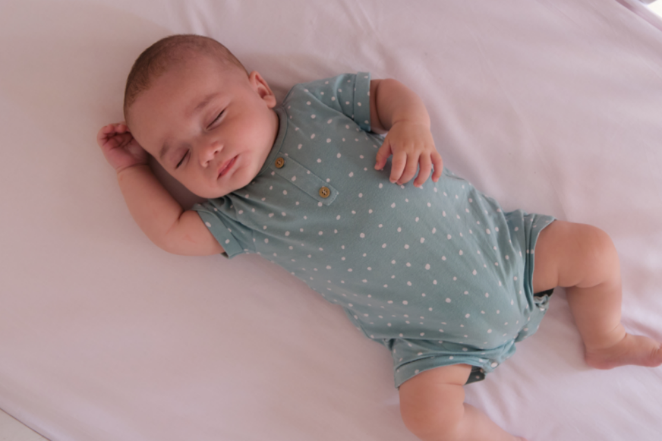 A baby sleeping safely on their back in an empty cot. They have one arm by their head, and another on their stomach. They are wearing a blue, polka-dot baby-grow.