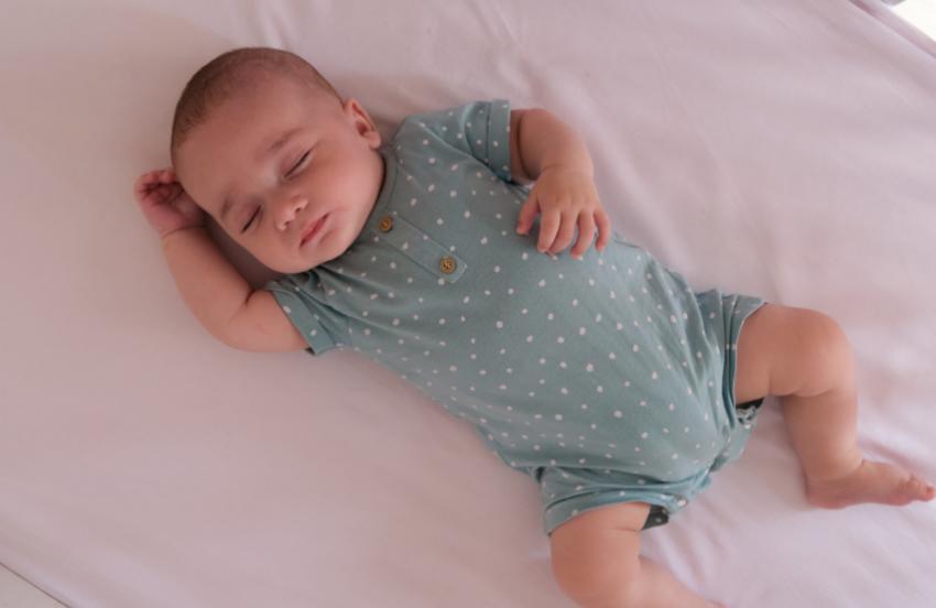 A baby sleeping safely on their back in an empty cot. They have one arm by their head, and another on their stomach. They are wearing a blue, polka-dot baby-grow.