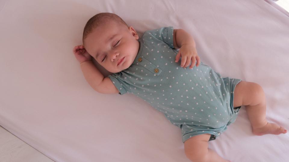 A baby sleeping safely on their back in an empty cot. They have one arm by their head, and another on their stomach. They are wearing a blue, polka-dot baby-grow.