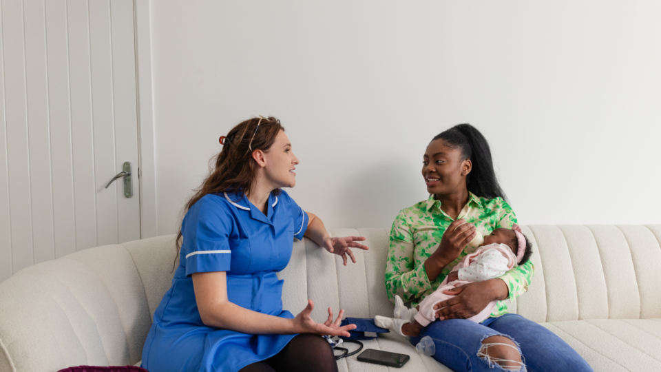 Mother bottle-feeding a baby, sitting on a sofa beside a health professional who is talking to her.