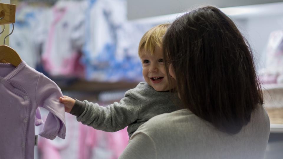 Baby and a mum are shopping together. The baby is smiling at someone out of frame and the mum is looking at the baby. The baby has blonde hair, light skin and is grasping onto a baby grow.