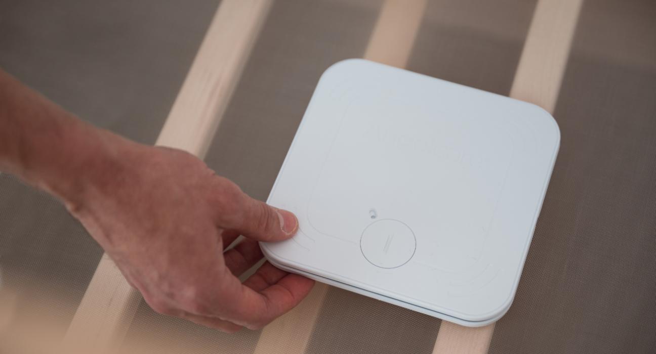 A hand is placing a monitor beneath a baby mattress in a cot